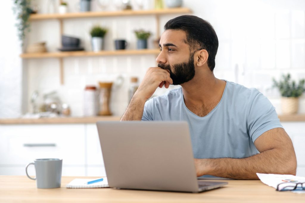 Thoughtful handsome indian or arabian man in casual t-shirt, freelancer, designer, working from home